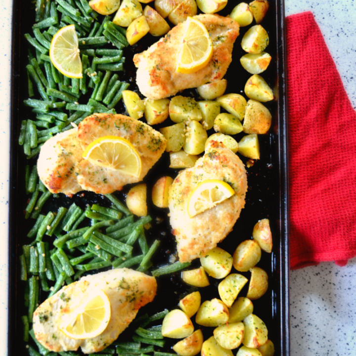 A baking sheet with four Sheet Pan Parmesan Garlic Chicken breasts topped with lemon, roasted potato chunks, and green beans, arranged on a speckled countertop with a red cloth beside the tray.