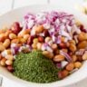 Close up of a white bowl on a white wood background with mixed beans, minced cilantro and red onion in it.