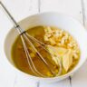 Close up of a white bowl with the dressing ingredients for mixed bean salad: Garlic, olive oil, dijon mustard, honey and white wine vinegar with a whisk in the bowl.