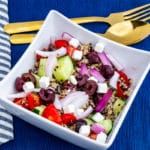 Close up of square white bowl full of colourful greek quinoa salad on blue placemat with blue and white striped napkin and gold cutlery