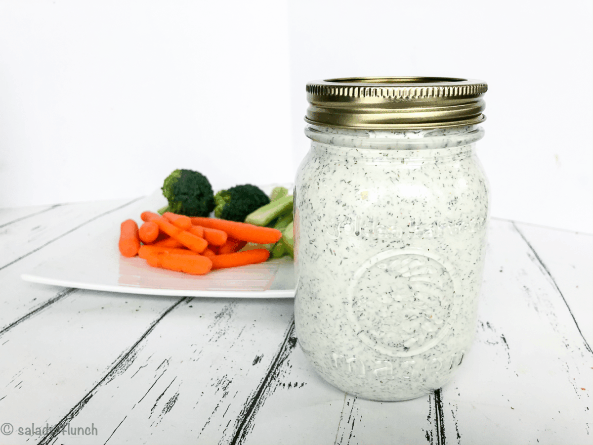 Close up of a mason jar full of vegan creamy dill dip with plate of carrots and broccoli in the background.