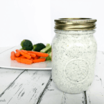 Close up of a mason jar full of vegan creamy dill dip with plate of carrots and broccoli in the background.
