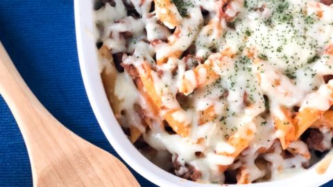 Penne pasta with hearty meat sauce with melted mozzarella cheese in a white casserole dish on a blue background
