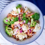 white bowl of broccoli and caulflower florets with chopped red onion, bacon bits, and matchstick carrots
