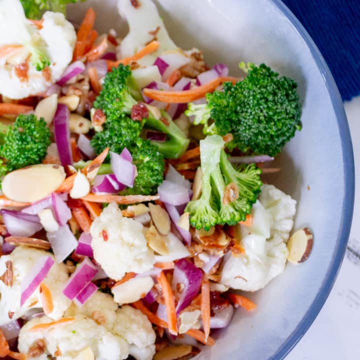 Close up of broccoli cauliflower salad with almonds, carrots and bacon in a white bowl with blue trim on a blue placemat with text overlay"Broccoli Cauliflower Salad with Bacon and Almonds"