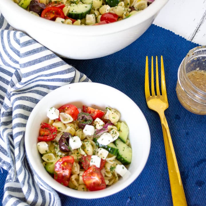 Large white bowl and smaller white bowl of colourful greek pasta salad with a small jar of greek dressing on a blue placemat