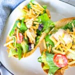 Two crispy edible taco bowls with chipotle chicken salad in them on a square white plate with black trim and a grey napkin beside the plate.