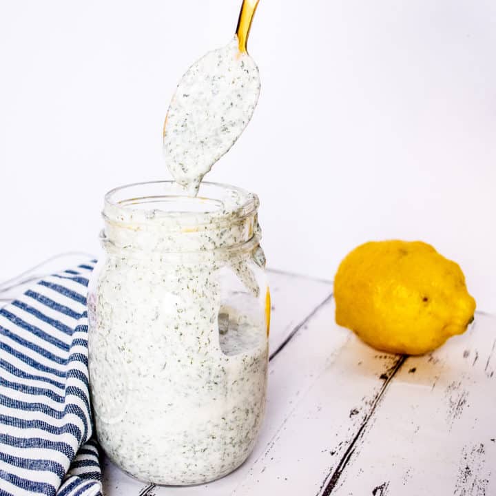 Gold spoon drizzling ranch dressing into a glass mason jar of creamy white ranch dressing on a white washed wood board. A blue and white striped napkin and lemon can be seen in the background.