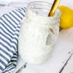 Gold spoon drizzling ranch dressing into a glass mason jar of creamy white ranch dressing on a white washed wood board. A blue and white striped napkin and lemon can be seen in the background.
