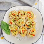 Close up of nine chickpea pesto avocado vegetarian pinwheel sandwiches on a square white plate with black trim.
