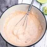 Close up of Chipotle Mayo in white bowl with black rim on a grey napkin with three lime slices in the background.