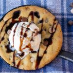 Close up of Salted Caramel and Chocolate Chip SKillet Cookie topped with vanilla ice cream and drizzled with chocolate on a blue and white plaid napkin.