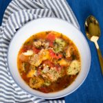 Close up of italian sausage and tortellini soup in a white bowl on a blue and white striped napkin.