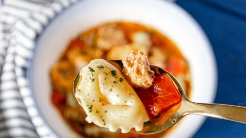 Close up of italian sausage and tortellini soup on a spoon with a white bowl of soup on a blue and white striped napkin in the background.