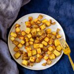 Top view of easy air fried home fries on a white plate on a blue napkin with a gold fork to the right of the plate.
