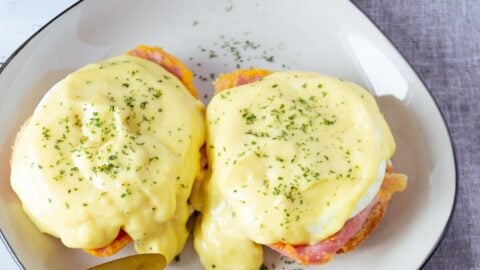 Close up photo of two delicious eggs benedict on a white plate with black trim on a grey napkin with a gold fork and knife.