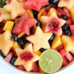 Overhead close up photo of a beautiful fruit salad with watermelon and peaches cut into stars, mango and blueberries in a white bowl.