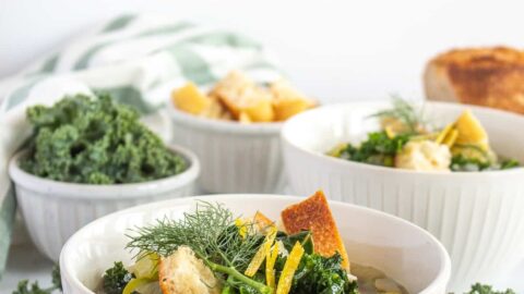 Side view of vegan spring minestrone soup garnished by two croutons in a white bowl with a bunch of fresh ale in the background.