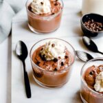 Close up of Vegan Chocolate Protein Pudding in a small glass bowl topped with coconut cream on a white counter with a black spoon and grey napkin.