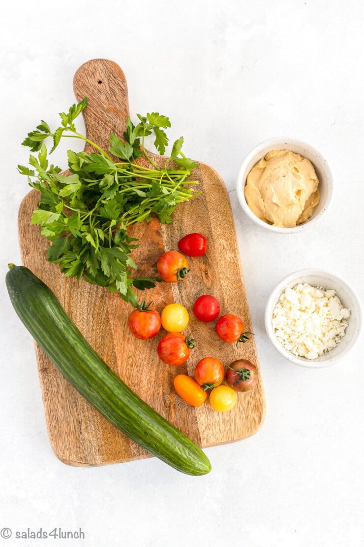 Cucumber, cherry tomatoes, hummus, parsley, and feta on a cutting board.