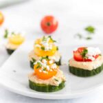 Close up photo of a square white plate withcucumber slices layered with hummus, a sliced cherry tomato topped off with feta and fresh parsley.