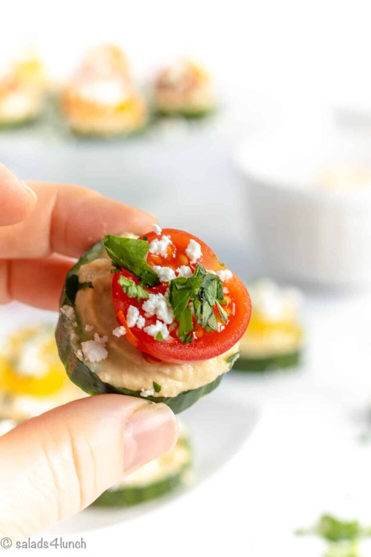 Close up photo of a hand holding a cucumber slice layered with hummus, a sliced cherry tomato topped off with feta and fresh parsley.
