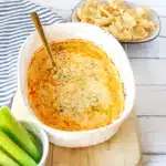 Close up of a white Corningware baking dish with buffalo chicken dip in it, sprinkled with parsley, and a bowl of tostitos and celery beside it.
