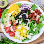 Close up of a white plate with southwest chopped salad on a wooden cutting board showing a cut avocado in the background.