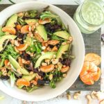 a white bowl with spring salad, mandarin oranges and cashews on a cutting board besid a mason jar with green goddess salad dressing.