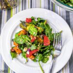 Overhead photo of a colorful vegetable salad with roasted red pepper, red onion, brussel sprouts, and sweet potato on a bed of baby spinach on a white plate.