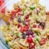 Bow Tie Greek Pasta Salad - Bow Tie Greek Pasta Salad 13 Close up of a glass bowl with cucumber, kalamata olives, tomatoes, red onion, feta cheese, parsley, and red bell pepper mixed together with bow tie pasta.