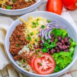 Image of a Low Sodium Ground Turkey Burrito Bowl Salad in a white bowl. The bowl is filled with a colorful mixture of cooked brown rice, seasoned ground turkey, black beans, diced tomatoes, sliced red onions, shredded lettuce, and taco blend cheese. A dollop of guacamole and a sprinkling of fresh cilantro top the vibrant ingredients.