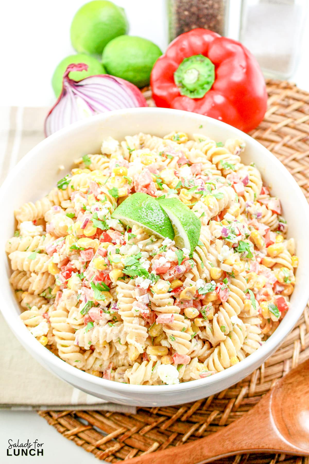 Mexican Street Corn Pasta Salad in a white bowl, garnished with a lime wedge, with red bell pepper, red onion, and limes in the background.