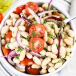 Close up of an italian white bean salad showing cannellini beans, red onions, cherry tomatoes and parsely mixed together in a white bowl.