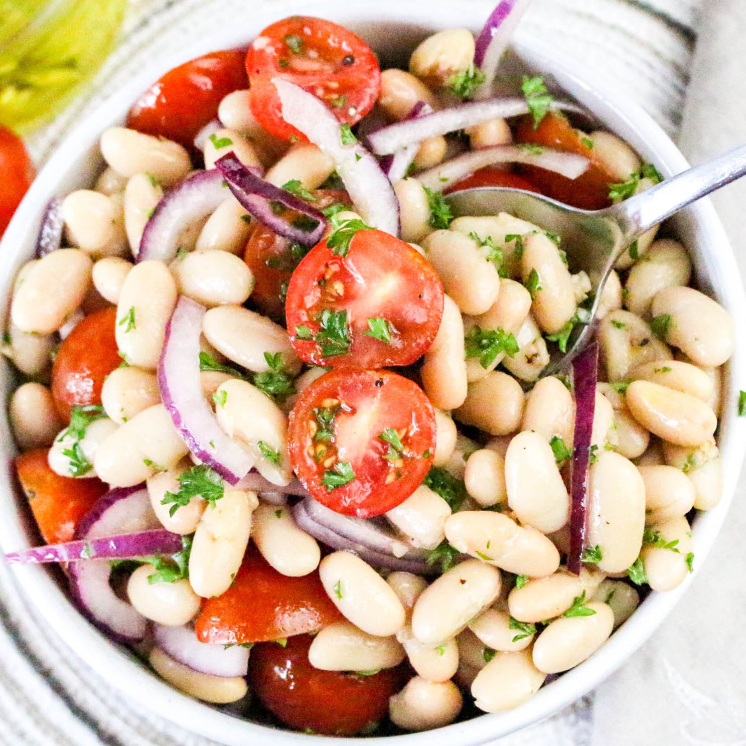 Close up of an italian white bean salad showing cannellini beans, red onions, cherry tomatoes and parsely mixed together in a white bowl.