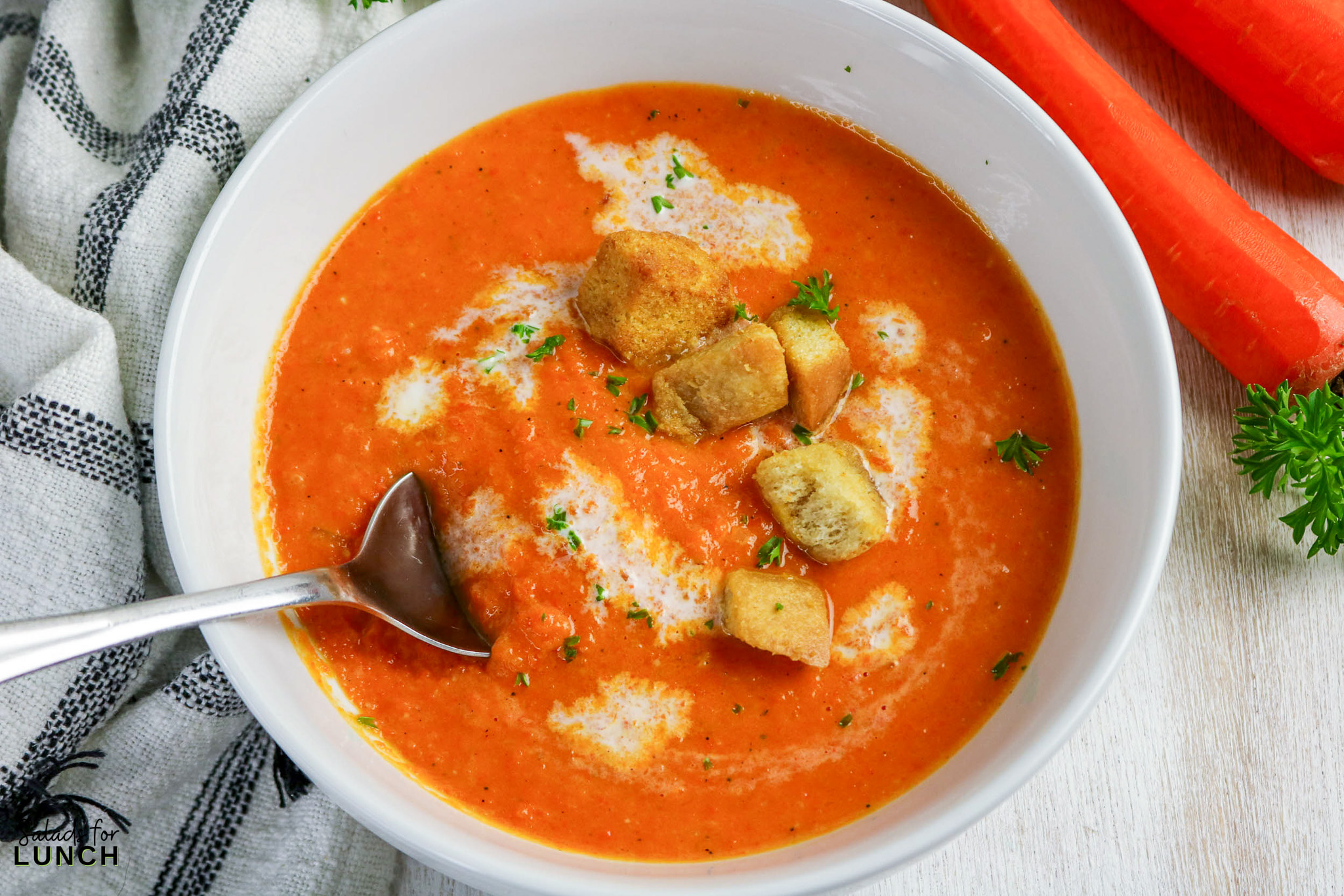 Creamy carrot soup in a white bowl, topped with croutons and parsley. Carrots on the side.