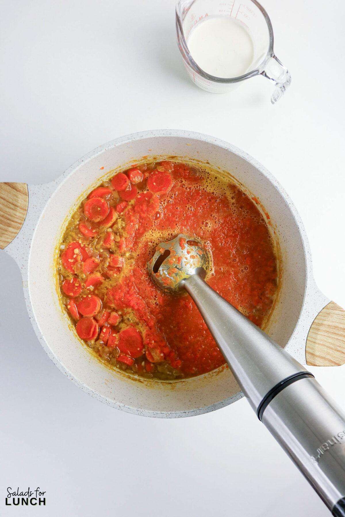 Creamy carrot soup being blended with an immersion blender, milk pitcher nearby.