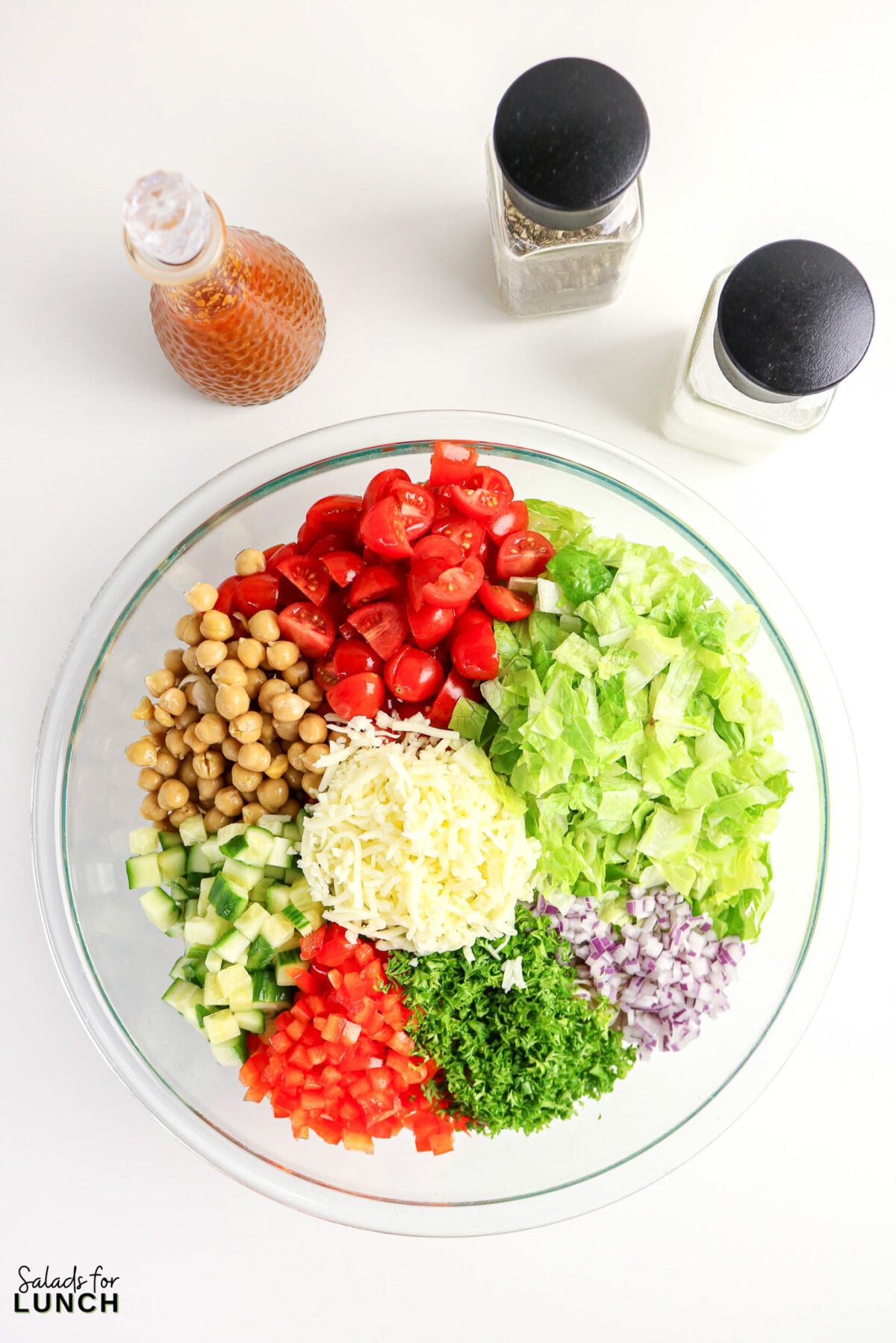 Chopped salad ingredients in a clear bowl with dressing and shakers