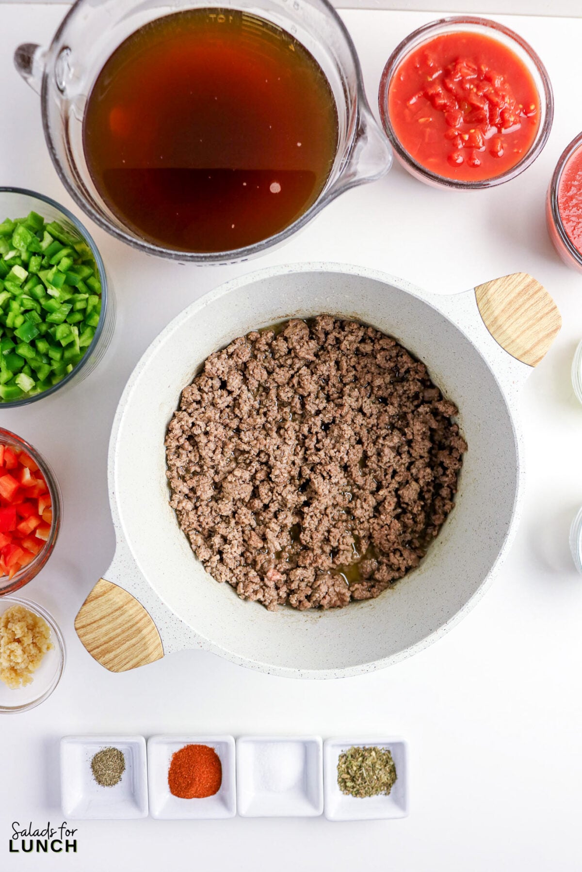 Ground beef in pot with diced vegetables, broth, and spices nearby.