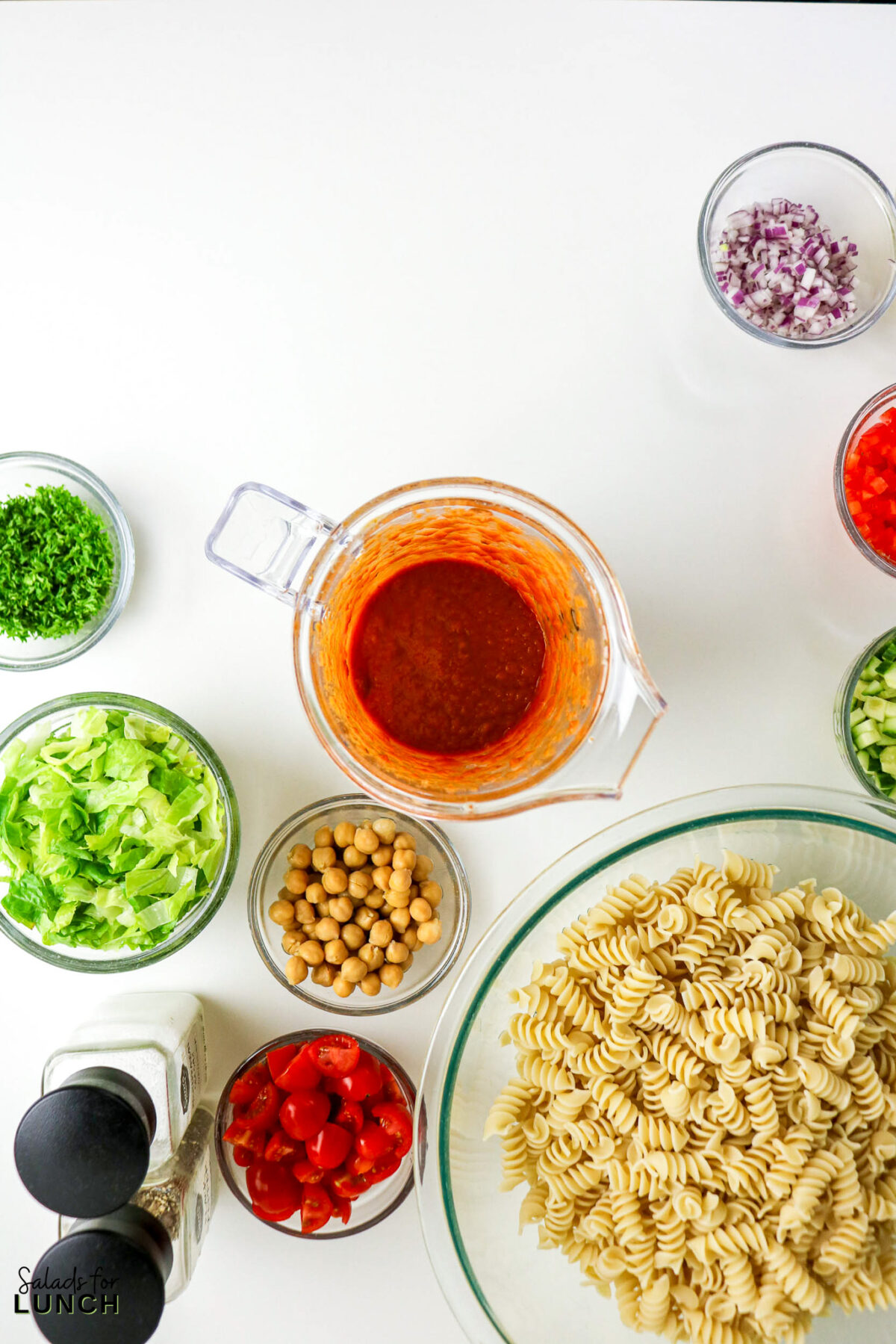 Ingredients for pasta salad: rotini pasta, chopped lettuce, chickpeas, tomatoes, red onion, parsley, and dressing.