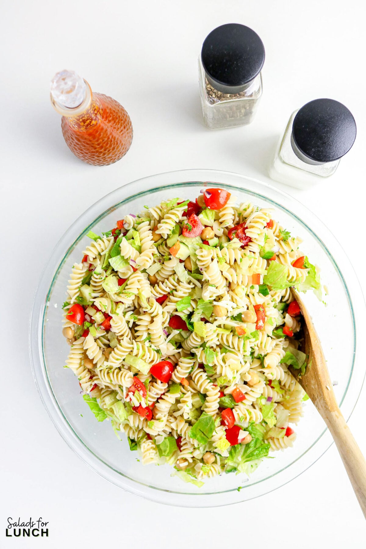 Rotini pasta salad with chickpeas, tomatoes, lettuce, and red onion in a glass bowl.