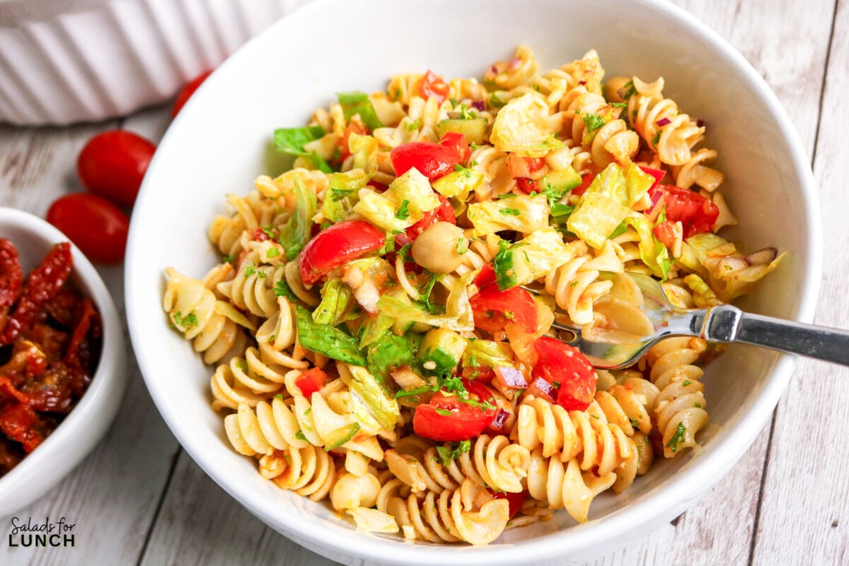 Rotini pasta salad with cherry tomatoes, cucumber, and lettuce in a white bowl.