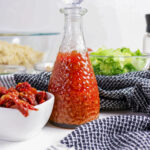 Glass bottle with red dressing, bowl of sun-dried tomatoes, and salad greens.