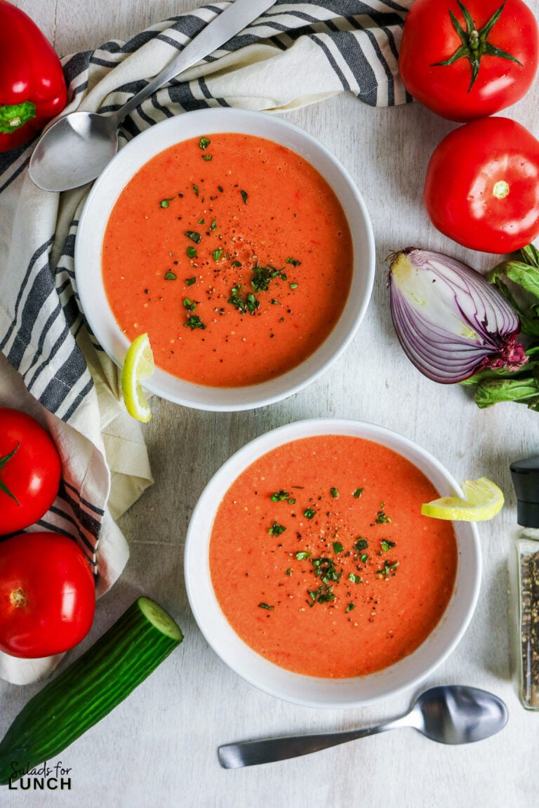 Two bowls of fresh gazpacho soup topped with herbs and lemon slices, surrounded by tomatoes and cucumber.
