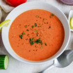 Bowl of gazpacho soup garnished with herbs and lemon slice, with fresh tomatoes and cucumber.
