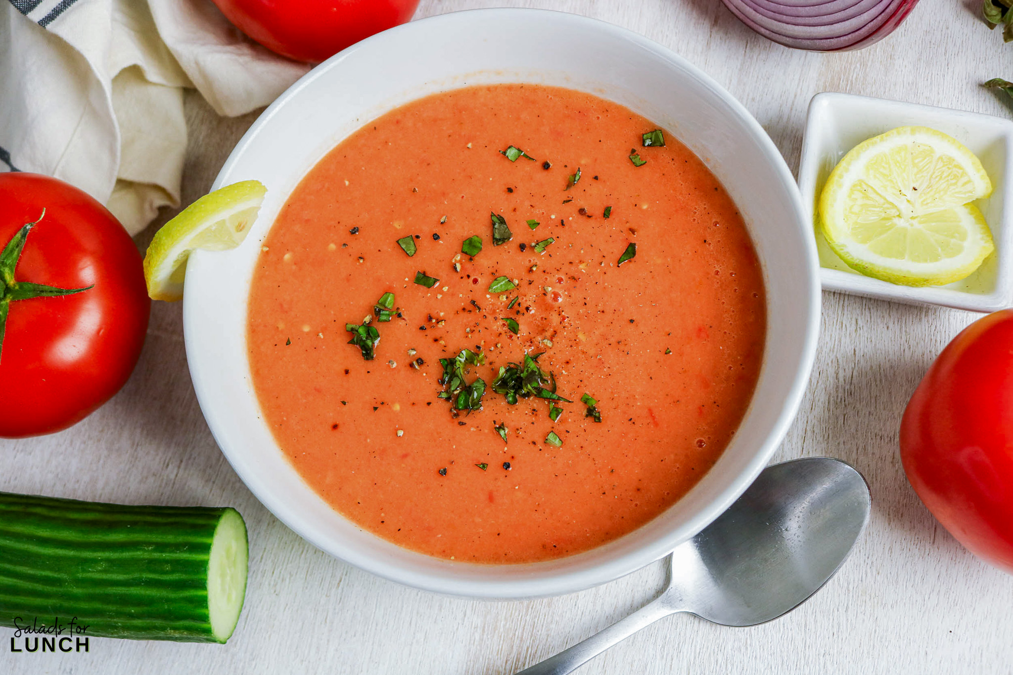 Bowl of gazpacho soup garnished with herbs and lemon slice, with fresh tomatoes and cucumber.