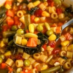 Close-up of a spoon scooping hearty vegetable soup with pasta, corn, peas, and greens.