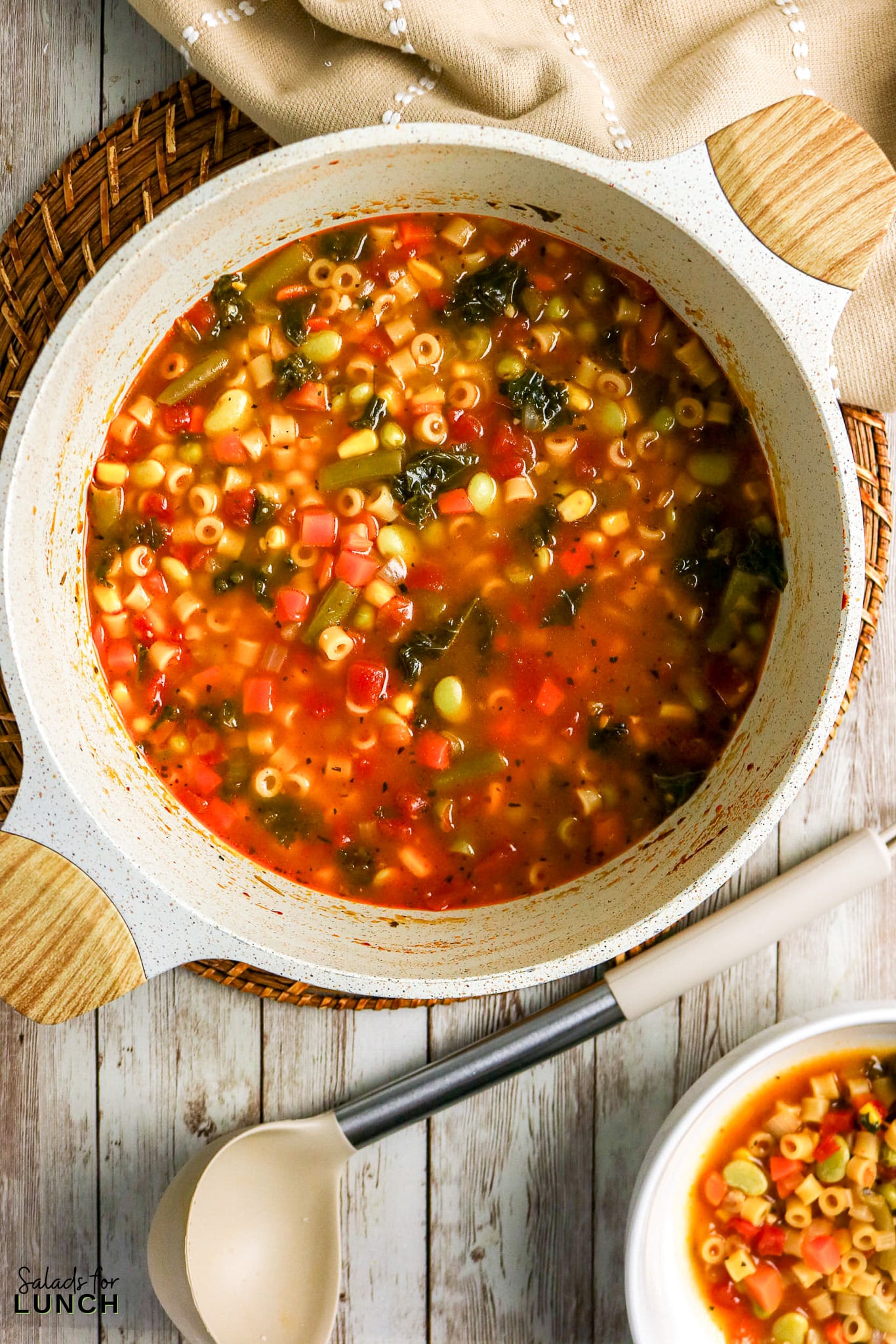 Hearty vegetable pasta soup in a white pot with wooden handles, served with a ladle.