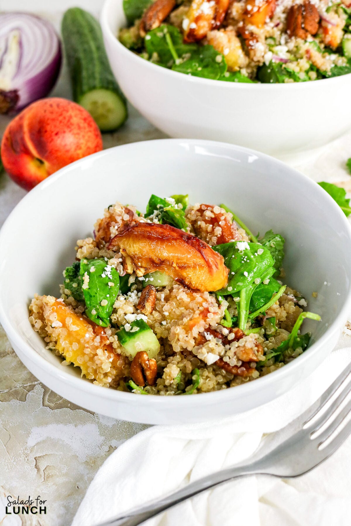 Bowl of grilled peach quinoa salad with arugula, toasted pecans, cucumber, and crumbled feta, with fresh peaches and ingredients in the background.