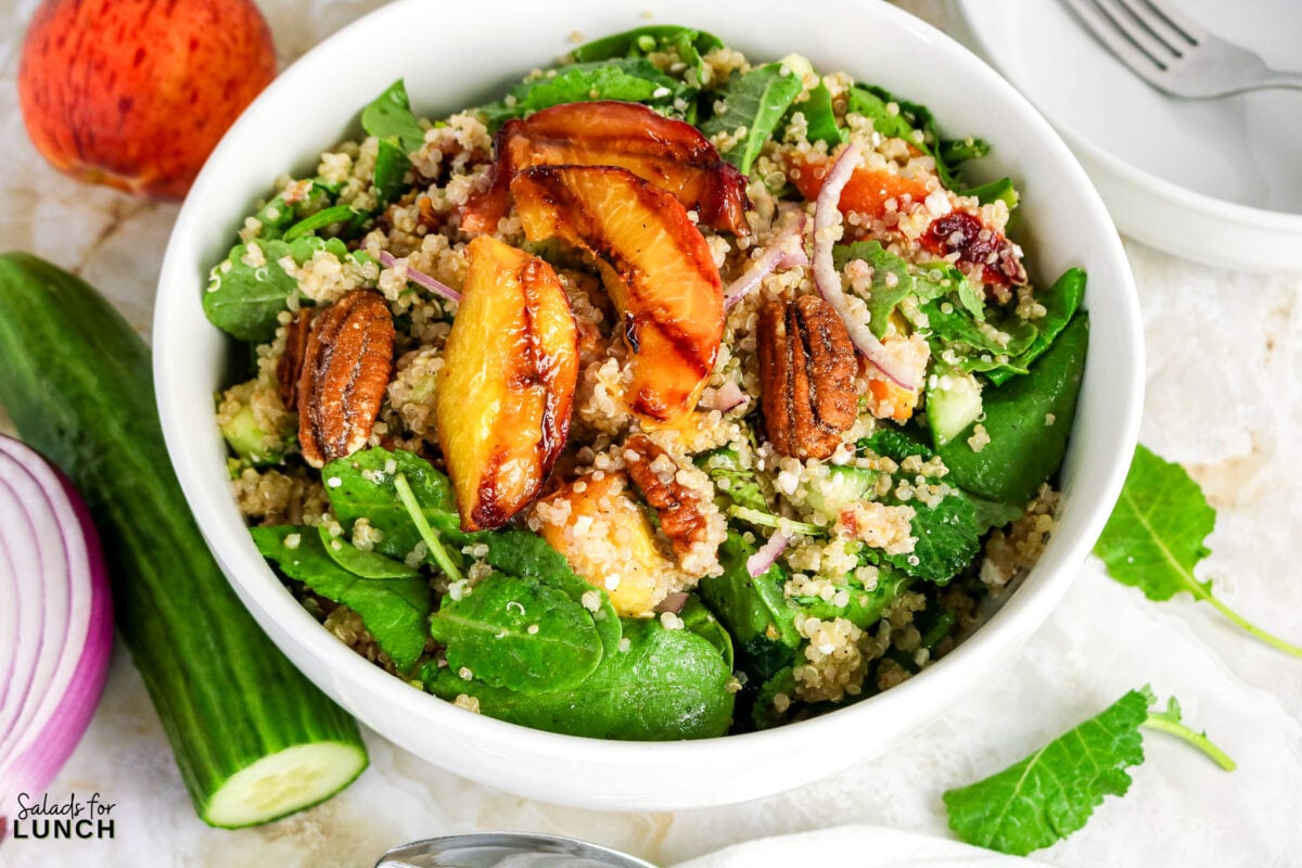 Quinoa salad bowl with grilled peach slices, candied pecans, spinach, kale, cucumber, and red onion.